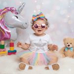Adorable baby girl, about one year old, wearing a rainbow tutu, white star-print bodysuit, glitter sneakers, and pink round sunglasses with a matching rainbow bow headband, seated on a white fuzzy rug next to a large unicorn balloon and a teddy bear, with a soft, bokeh background.