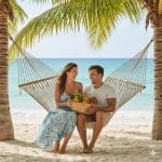 Smiling couple sitting together on a hammock between palm trees at a tropical beach, holding a basket of fresh fruit with turquoise ocean in the background