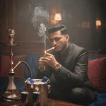 A young man in a white shirt and black pants sits with his eyes closed, leaning against a bare-branched tree trunk in a lush, dense green field under a diffused, cloudy sky.