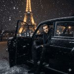 Stylish man in all-black winter outfit sitting in a classic car during snowfall with the illuminated Eiffel Tower glowing in the background, Paris at night