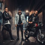 A group of three stylish men in a dimly lit urban alleyway at night. One man leans against a brick wall, another stands in the center with sunglasses and a denim jacket, and the third sits on a vintage motorcycle. They are all wearing black, modern, and edgy clothing.