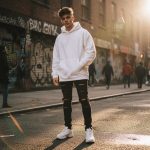 Ultra-realistic 8K image of a young modern boy standing on a city street, wearing a white oversized hoodie, black ripped jeans, and white sneakers, with a messy undercut hairstyle. Graffiti-covered walls in the background and golden hour sunlight reflecting on the pavement create a cinematic urban vibe.