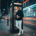 This urban cinematic editorial portrait from the “London Light Trails Series” captures a stylish young man standing near St. Paul’s Cathedral on a cool London night. Dressed in a black hoodie, white cargo pants, and white sneakers, he exudes effortless confidence while holding a Starbucks coffee cup. The glowing vintage telephone booth bathes him in a warm orange light, contrasting beautifully with the cool city blues and dynamic red and white car light trails passing by. Shot on a DSLR camera (f/1.8–2.2) for a dreamy shallow depth of field, this image blends modern street fashion with cinematic storytelling — a perfect fusion of calm style and urban energy.