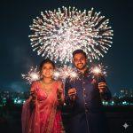 A young Indian couple celebrating Diwali outdoors at night, holding glowing sparklers with colorful fireworks lighting up the sky behind them. The woman wears a pink lehenga and the man a navy blue kurta, both smiling joyfully in elegant festive attire. Captured in ultra-realistic 8K photography with warm lights and natural skin tones.
