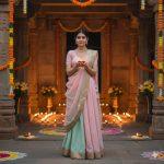 A beautiful young Indian woman wearing a simple pastel lehenga with golden borders, standing in a temple courtyard decorated with lamps and flowers, holding a glowing diya in her hands, with soft warm light illuminating her calm and spiritual face, captured in realistic natural photography.