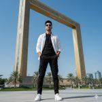 Ultra-realistic cinematic AI portrait of a modern young man posing confidently near the Dubai Frame, wearing a monochrome outfit with black cargos, a white shirt, and sneakers under a clear blue sky and geometric background.