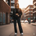 A stylish young woman stands confidently in an urban city setting, wearing high-waist jeans, a crop top, a leather jacket, and trendy sneakers. Her sleek, shoulder-length hair with subtle highlights glows under soft golden-hour lighting. The background features modern architecture, graffiti walls, and blurred bokeh lights, captured in ultra-detailed 8K resolution with cinematic lighting.