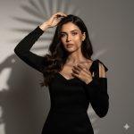 Ultra-realistic 12K cinematic portrait of a young woman with long dark wavy hair standing near a softly lit window, wearing a black off-shoulder dress and delicate necklace, illuminated by warm natural sunlight.