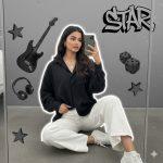 Ultra-realistic 8K cinematic portrait of a stylish young woman sitting casually on the floor, taking a mirror selfie. She wears a loose black shirt and white pants, holding her shirt with one hand and the phone with the other. Minimal jewelry, relaxed expression, soft natural light, and a white outline glow. The background features a modern grayscale collage with an electric guitar, headphones, dice, cat face decor, 3D stars, and “STAR” graffiti text.