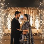 A bride in a glittering lehenga and a groom in a dark sherwani stand under sparkling fireworks at night. Soft illumination highlights their joyful expressions, with a slightly blurred background emphasizing the romantic glow and festive celebration.