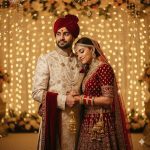 A cinematic Indian wedding portrait capturing a bride in a red lehenga with intricate golden embroidery, adorned with traditional jewelry, gently resting her head on the groom’s shoulder. The groom wears a cream sherwani with a red stole. They stand close together against a backdrop of warm golden fairy lights, creating a soft bokeh effect and a dreamy, romantic atmosphere in ultra-detailed 8K resolution.