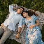 A romantic pre-wedding photoshoot during golden hour showing a couple holding hands and embracing in a lush green park. The woman wears a flowing white dress, gently lifted by a breeze, and the man wears a casual light shirt with grey shorts. The scene is illuminated by warm sunset light, creating a dreamy bokeh effect with scattered petals and distant string lights.