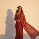 A woman wearing a flowing plain red saree with a matching sleeveless blouse, a white flower tucked in her dark wavy hair, standing against a plain white background. Golden hour sunlight casts a warm glow and dramatic shadow, highlighting her serene expression as she looks slightly off-camera.