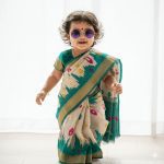 Cute Indian baby girl walking in a modern stylish saree, curly hair bouncing, wearing tiny trendy goggles, smiling playfully in soft natural light. Ultra-realistic 4K photography style with vibrant colors.